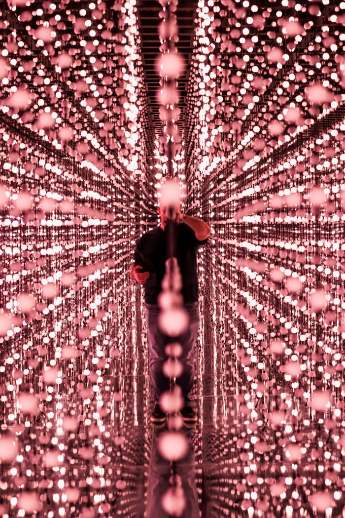 gallery-2 A person reflected in an infinity mirror room filled with pink LED lights, creating a stunning visual effect.